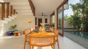 A modern dining area with a wooden table set for six, surrounded by wicker chairs, and a view of a swimming pool. - Villa Omah Bumbak (Badung)