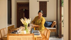 A person pouring a drink into a glass at a wooden table with a vase of flowers, a sofa, and a patterned wall hanging. - Villa Omah Bumbak (Badung)