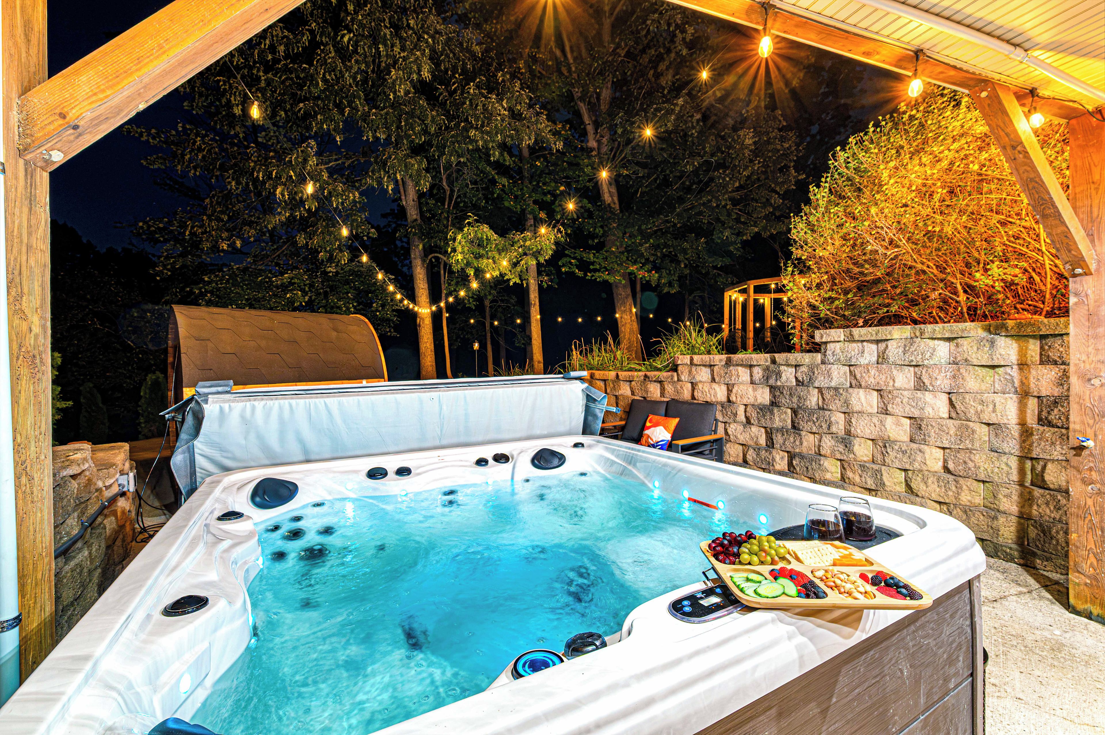 A hot tub with a wooden deck and a stone wall, surrounded by trees and string lights.