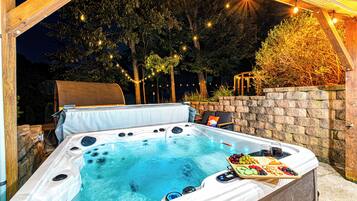 A hot tub with a wooden deck and a stone wall, surrounded by trees and string lights.