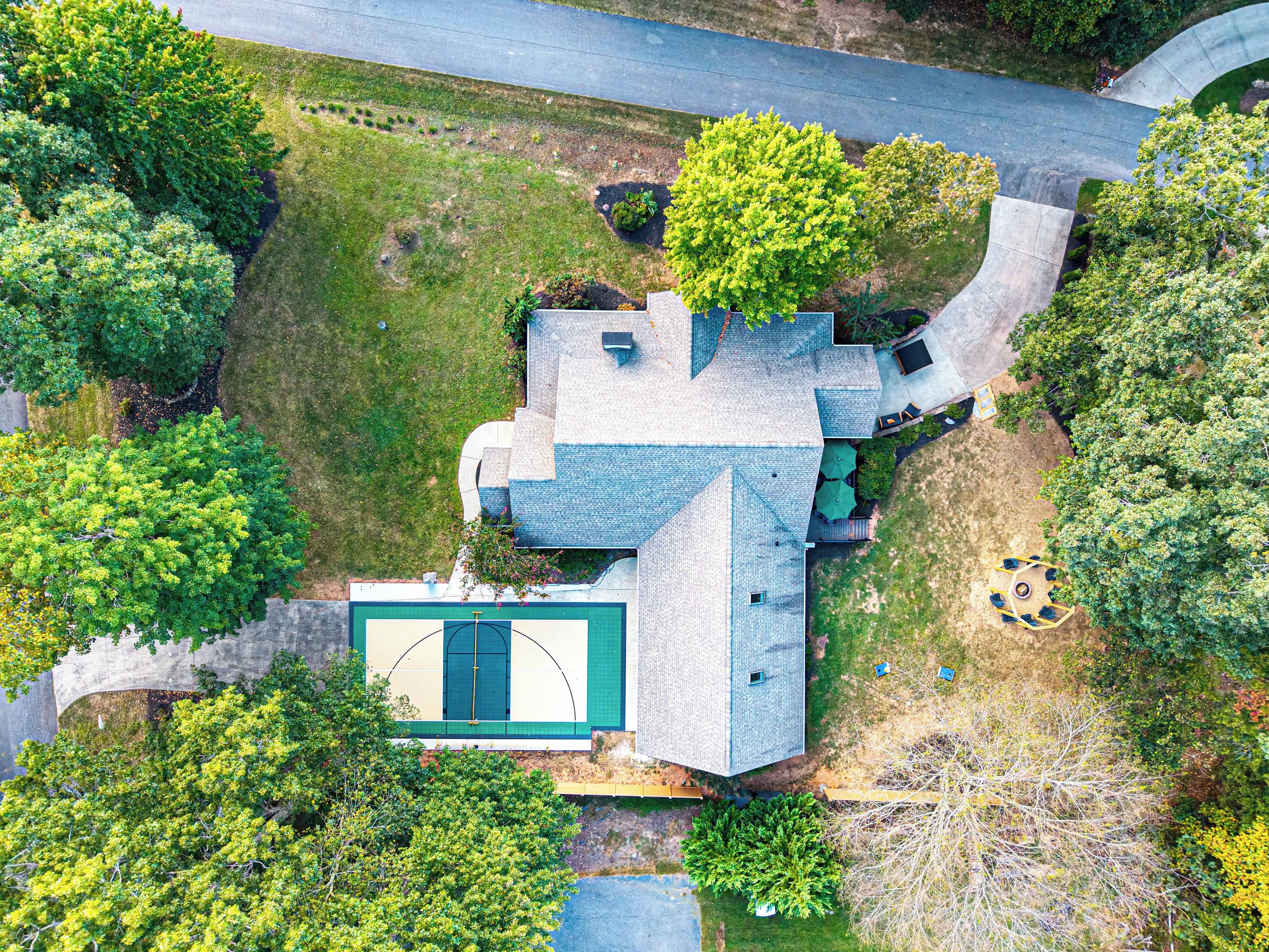 An aerial view of a residential property with a basketball court, a driveway, and surrounding greenery.