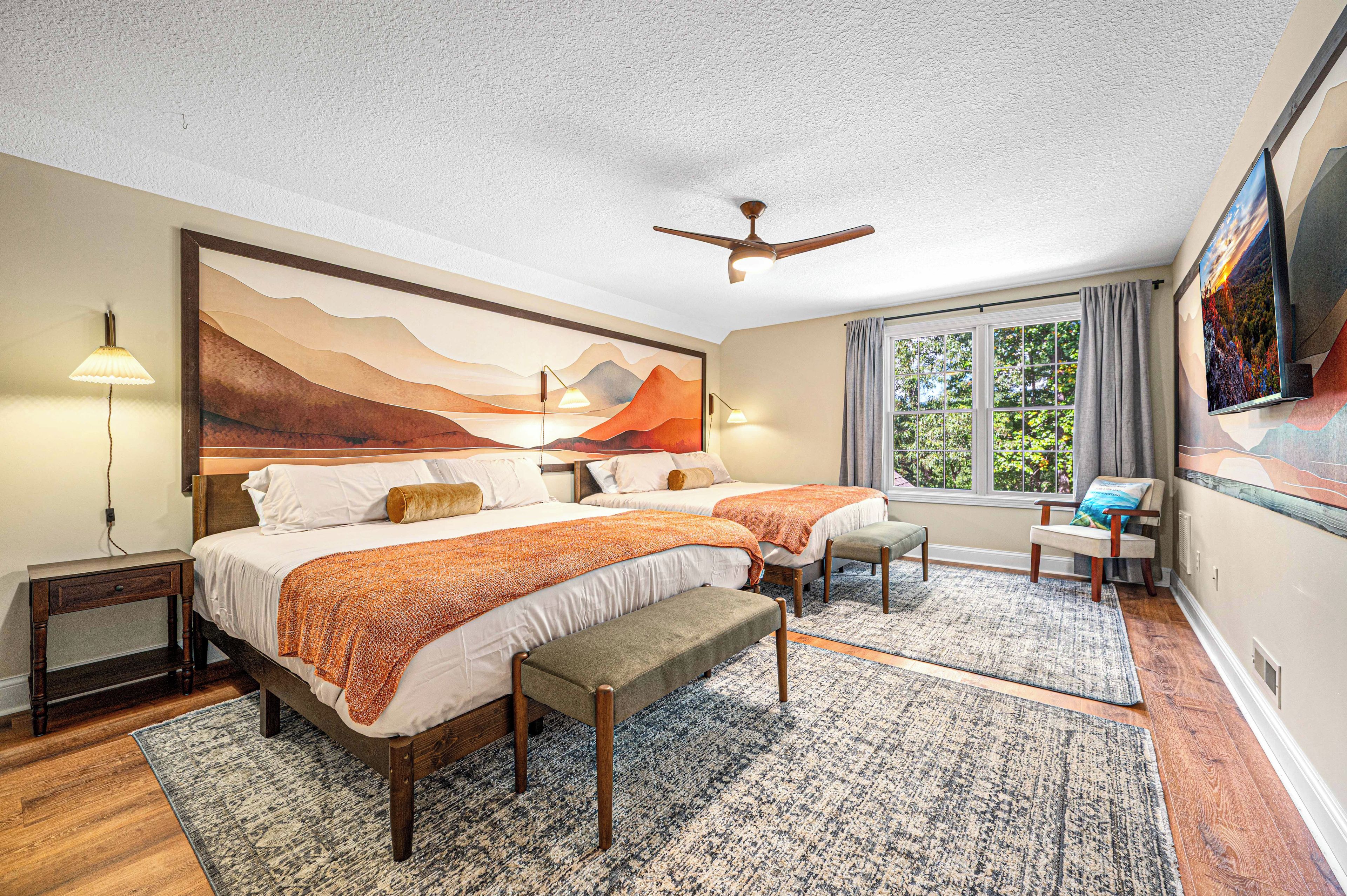 A bedroom with a large bed, two bedside tables, a bench, a ceiling fan, a window with curtains, and a painting on the wall.