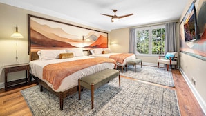 Quarto com uma cama grande, duas mesas de cabeceira, um banquinho, um ventilador de teto, uma janela com cortinas e um quadro na parede.