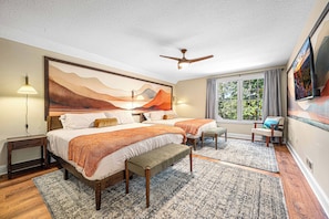 Une chambre avec un grand lit, deux tables de chevet, un banc, un ventilateur de plafond, une fenêtre avec des rideaux et un tableau au mur.