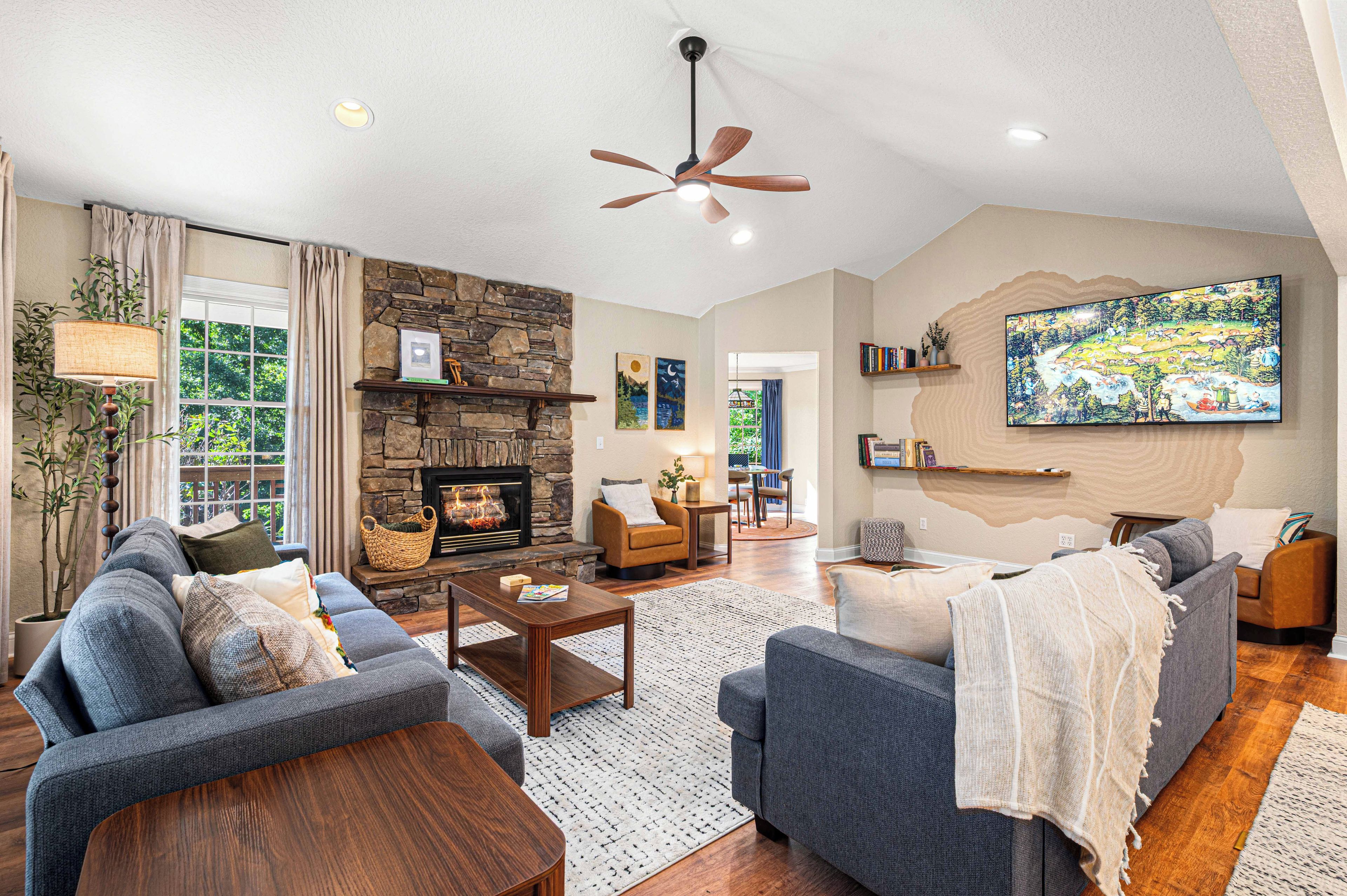 A spacious living room with a stone fireplace, comfortable seating, a ceiling fan, and a large painting on the wall.