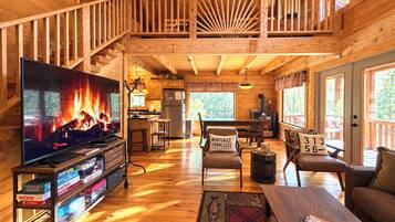 A spacious wooden cabin interior with a large television, a fireplace, a dining area, and a balcony.