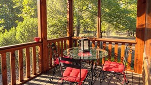 Een houten terras met een glazen tafel en stoelen, omgeven door een houten hekwerk, met uitzicht op een bebost gebied.