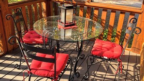 Een tafel met glazen blad, met een lantaarn, omringd door metalen stoelen met rode kussens, op een houten terras met uitzicht op bomen.