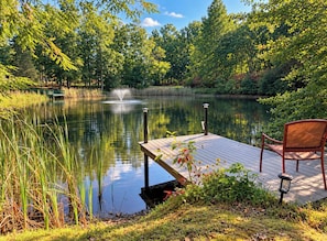 Escena tranquila junto al lago con un muelle de madera y una silla, rodeada de vegetación exuberante y un cielo despejado.