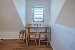 Ein Eckzimmer mit Holztisch und zwei Hocker, einem Fenster mit Jalousien und einer weißen Wand.