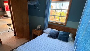 A bedroom with a bed, a nightstand, a window with curtains, and a door leading to a kitchenette. - perfect for family. Angler and nature guides (Owens Town)