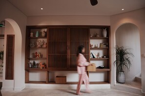 Une personne qui traverse une pièce en portant un panier, et dans laquelle on peut voir une étagère en bois où sont exposés divers objets décoratifs.