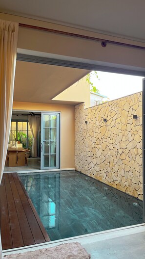 A modern outdoor pool area with a stone wall, a wooden deck, and a glass door leading to an adjacent room. - Serene Tropical Stay with Pool Near Hoi An Old Town (Đà N ẵng)