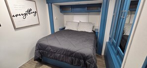 A bedroom with a bed, a nightstand, a wall-mounted shelf, and a wardrobe with blue doors. - Country Camping w/farm amimals (Brooksville)