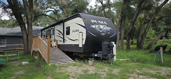 Un camper PUMA con un portico coperto, una piccola area cucina esterna e una strada asfaltata che conduce a un pontile di legno.