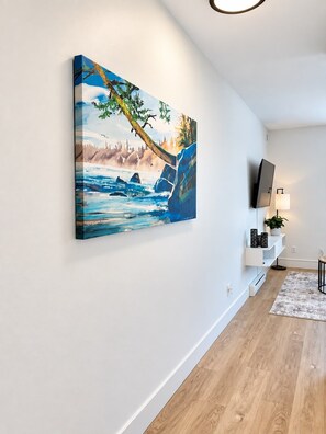 A room with a mounted painting of a landscape, a flat-screen TV, a white console table with a lamp, and a patterned rug. - The Haven Retreat | 1-Bedroom | Near Victoria (LANGFORD)