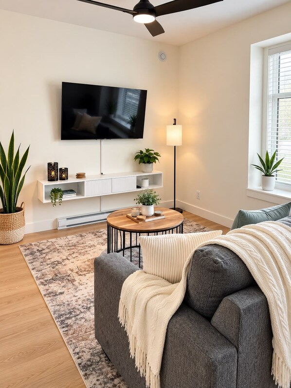 A modern living room with a flat-screen TV, a ceiling fan, a round coffee table, and a sofa with a throw blanket. - The Haven Retreat | 1-Bedroom | Near Victoria (LANGFORD)