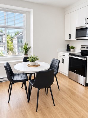 A modern kitchen with white cabinetry, a stainless steel stove, and a round dining table with black chairs. - The Haven Retreat | 1-Bedroom | Near Victoria (LANGFORD)