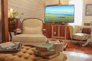 Een gezellige woonkamer met een aan de muur bevestigde televisie, een comfortabele fauteuil, een klein tafeltje met boeken en een decoratieve mand.