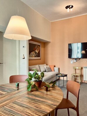 A modern living room with a sofa, a dining table with chairs, a TV mounted on the wall, and a ceiling light fixture. - New Flat with Artistic Flair (Milano)