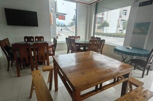 Un comedor con una mesa y sillas de madera, un televisor montado en la pared y ventanas amplias con vista a la calle.