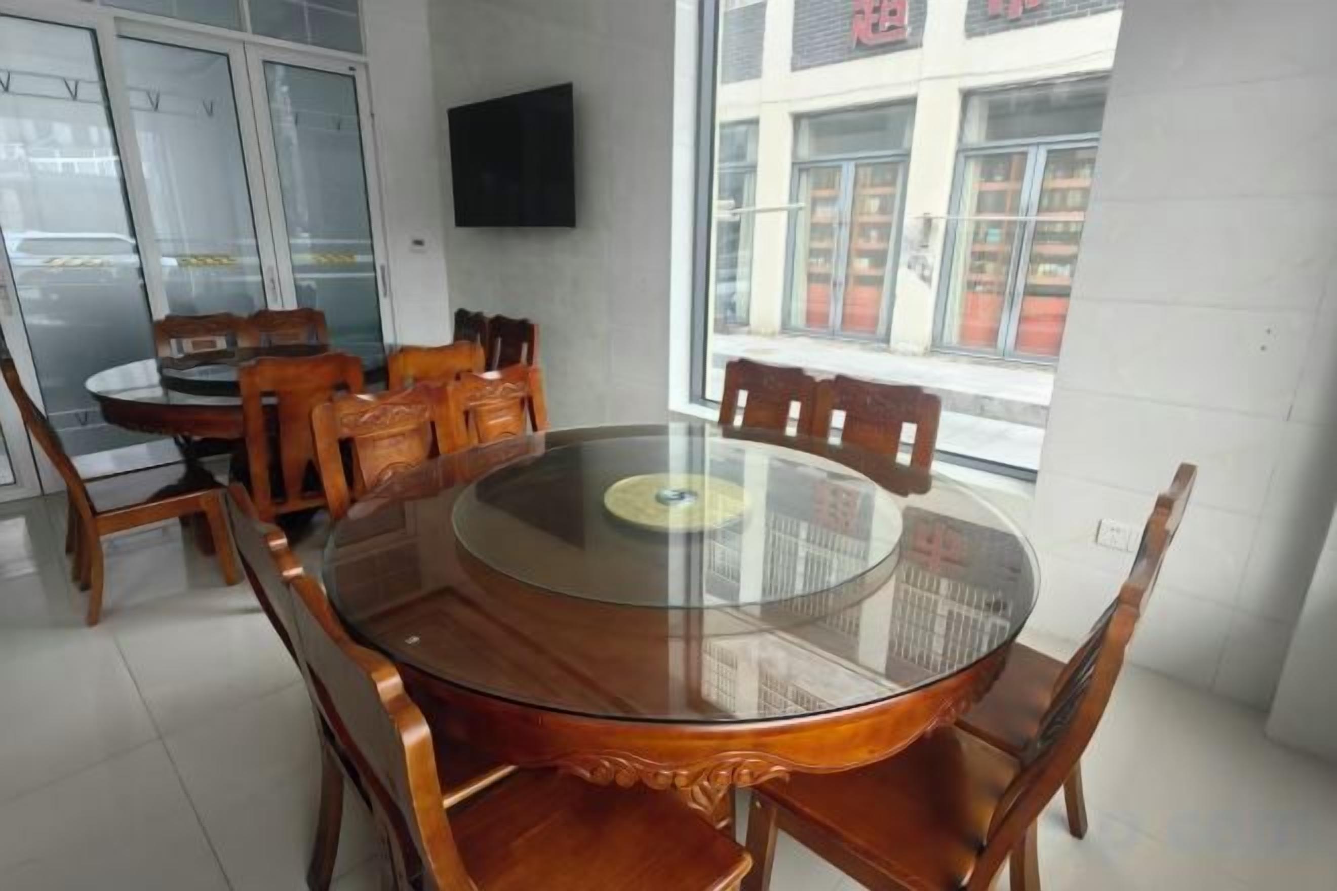 Un comedor con una mesa de vidrio redonda rodeada de sillas de madera, un televisor montado en la pared y una ventana con vistas a la calle.