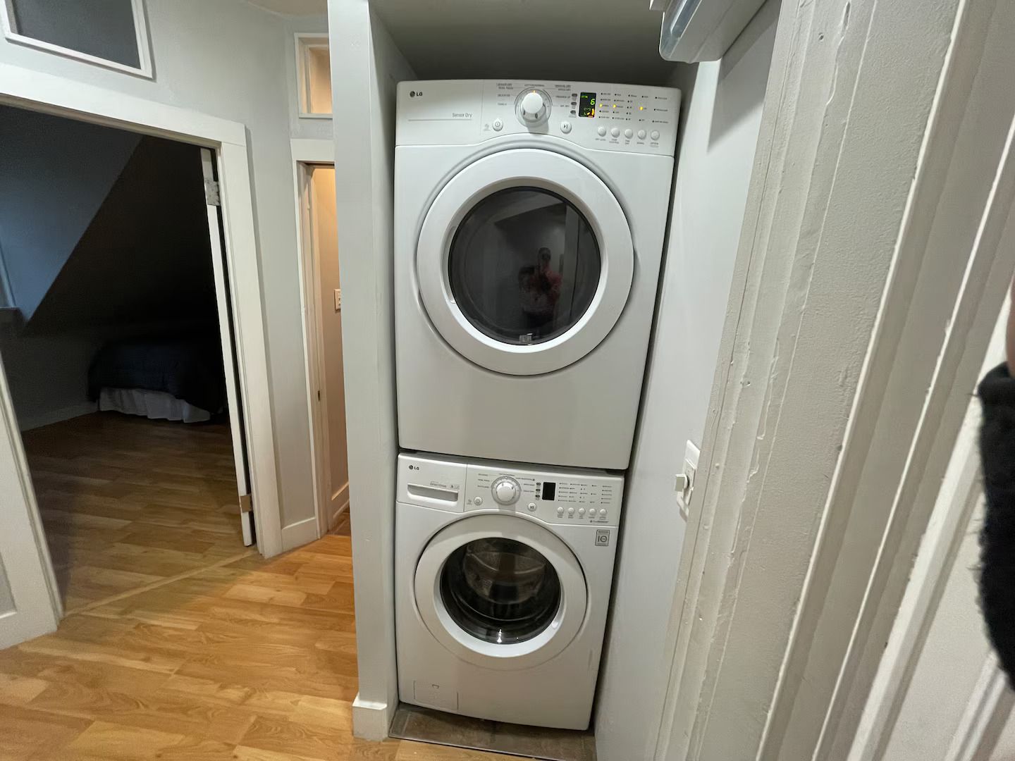 A laundry room with two stacked washing machines, a wooden floor, and a door leading to another room.
