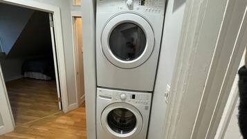 A laundry room with two stacked washing machines, a wooden floor, and a door leading to another room.