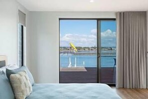 A bedroom with a large bed, a sliding glass door leading to a balcony with a view of the sea and a distant yellow structure. - SouthWind your premier escape destination in Merimbula (Merimbula)