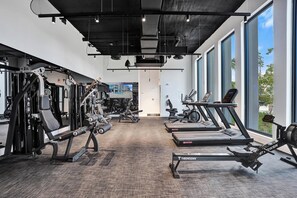 A gym with various exercise equipment, including treadmills and weight machines, with large windows and a view of the outdoors. - Oceanfront 3BR | Balcony, Pool & Beach Steps (Pompano Beach)