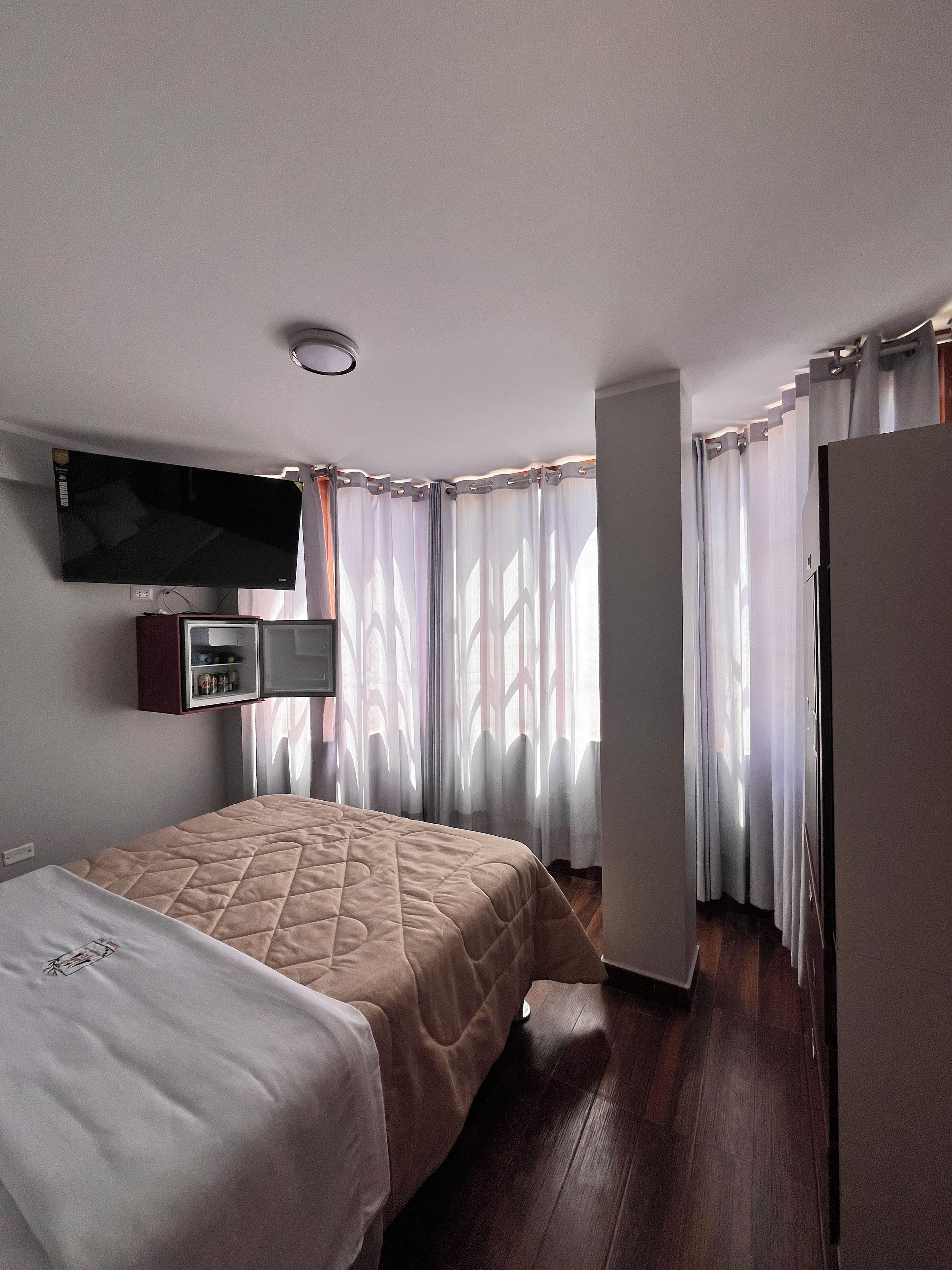 Habitación de hotel con cama, televisor de pantalla plana en la pared, armario y ventana con cortinas.