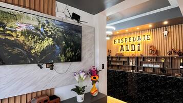 A modern hotel lobby with a large TV mounted on a wall, a reception desk with 'HOSPEDALE ALDI' signage, and a wooden floor.