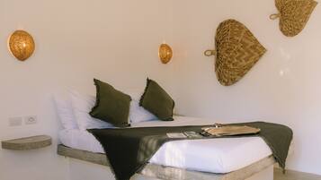 A bed with white bedding, two green pillows, and a dark throw blanket. There are two wall-mounted light fixtures and two woven wall hangings.