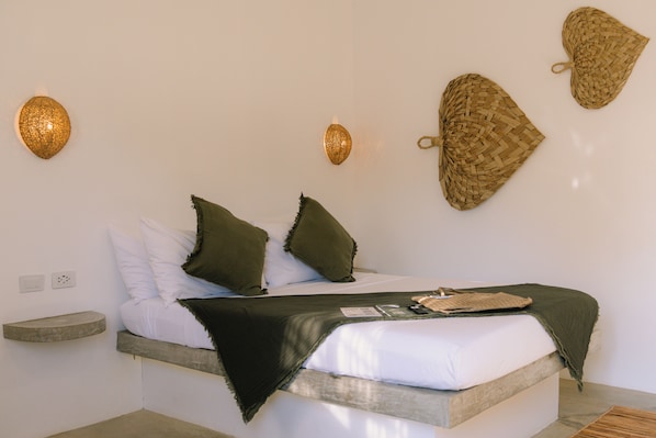 Una cama con ropa de cama blanca, dos almohadas verdes y una manta oscura. Hay dos lámparas de pared y dos tapices tejidos.
