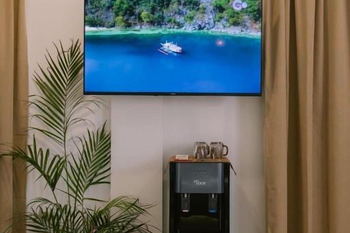 A flat-screen TV mounted on a wall, a water dispenser, and two potted plants in a room with beige curtains.