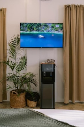 A flat-screen TV mounted on a wall, a water dispenser, and two potted plants in a room with beige curtains. - SAMPA El Nido (El Nido)