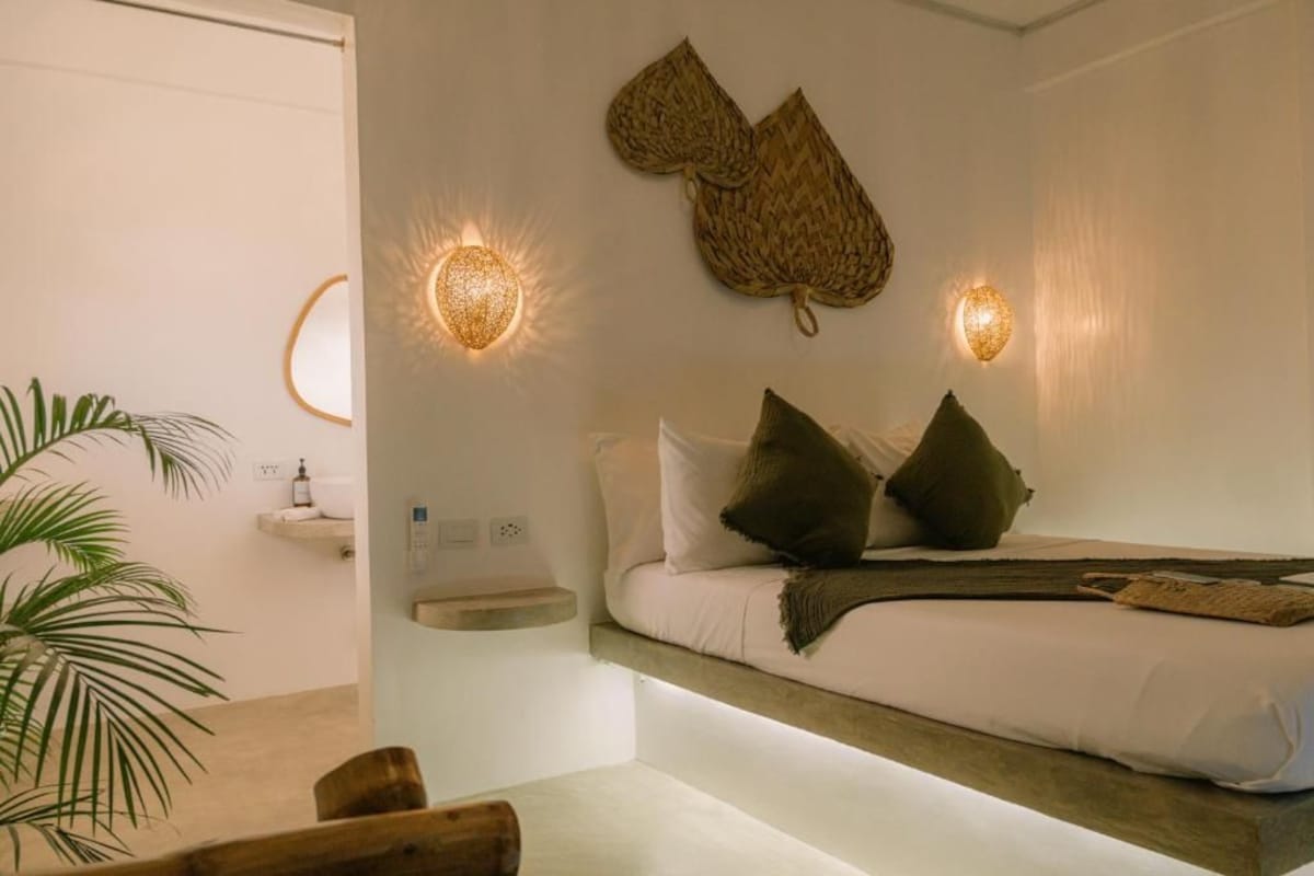A bedroom with a bed, pillows, a bedside table, a wall-mounted lamp, and a hanging woven basket.