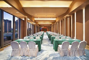 Un salón de conferencias con filas de mesas y sillas, ventanas amplias y vistas a un paisaje urbano.
