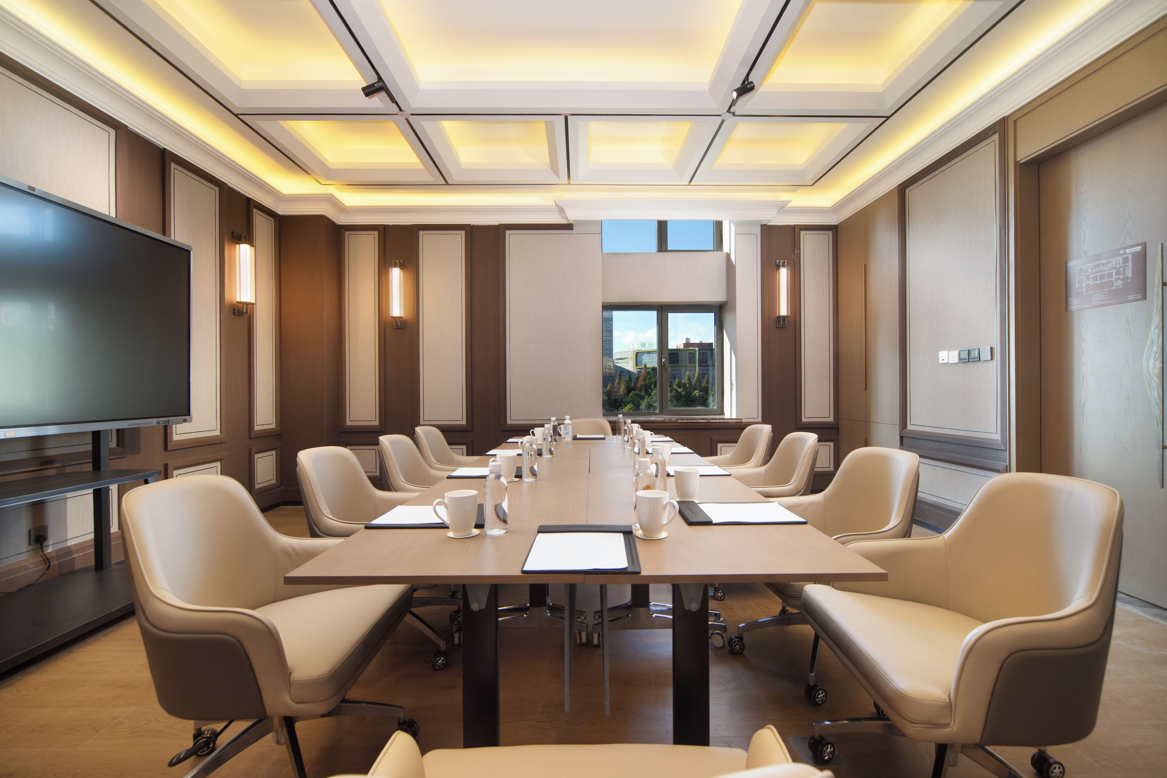 Un salón de conferencias con una larga mesa, sillas, una televisión y una ventana con vistas a edificios.