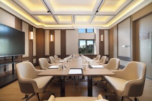 Un salón de conferencias con una larga mesa, sillas, una televisión y una ventana con vistas a edificios.