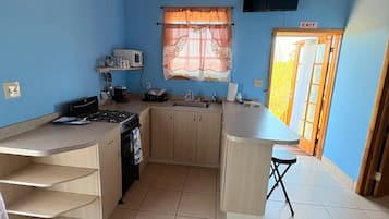 Une petite cuisine équipée d’une cuisinière, d’un évier et de placards, une fenêtre avec des rideaux et une porte marquée « SORTIE ».