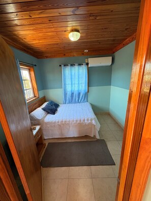 Ein Schlafzimmer mit Holzboden, einem Bett mit weißer Bettdecke, einem Fenster mit Vorhang und einer Klimaanlage.