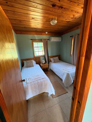Ein Zimmer mit zwei Betten, Holzboden, einem Fenster mit Vorhängen und einer Klimaanlage.