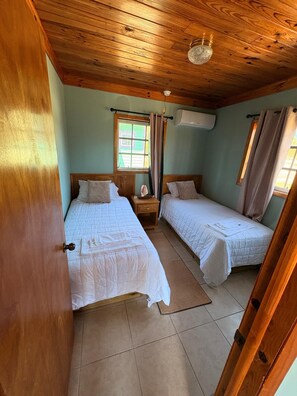 Ein Zimmer mit zwei Betten, Holzboden, Klimaanlage und einem Fenster mit Vorhängen.