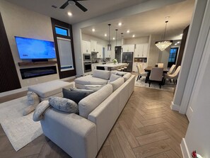 Een moderne woonkamer met een grote hoekbank, een flatscreen televisie en een eethoek met een kroonluchter.