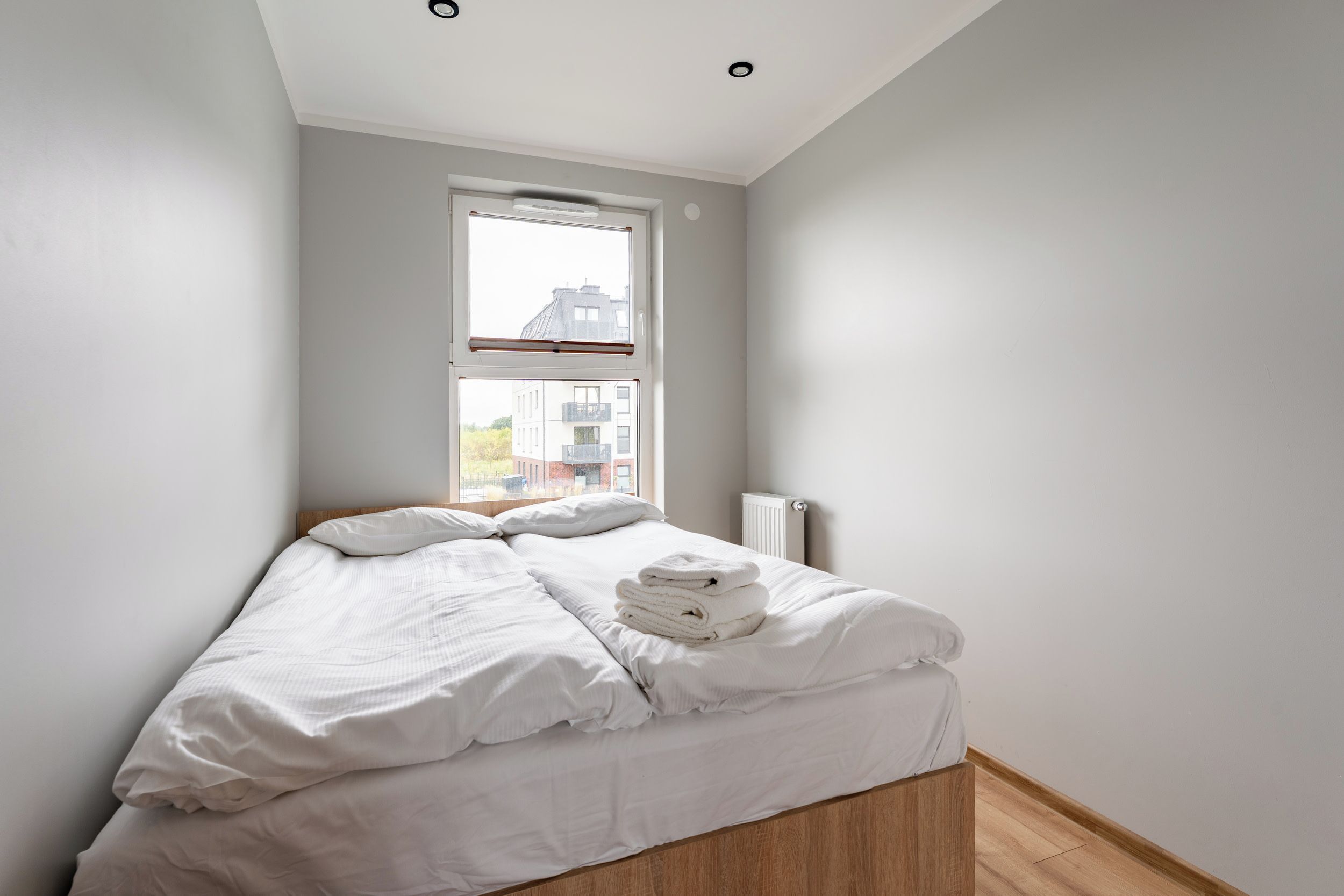 A bedroom with a bed, a window, a radiator, and a view of buildings outside.