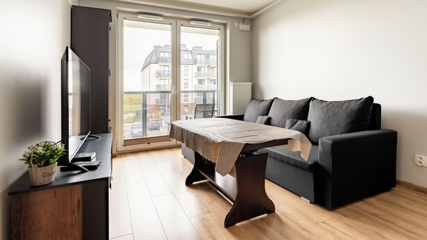 A modern living room with a sofa, a coffee table, a TV, and a view of the outside. - Gdańsk Koprowskiego Apartment #18 (Gdańsk)
