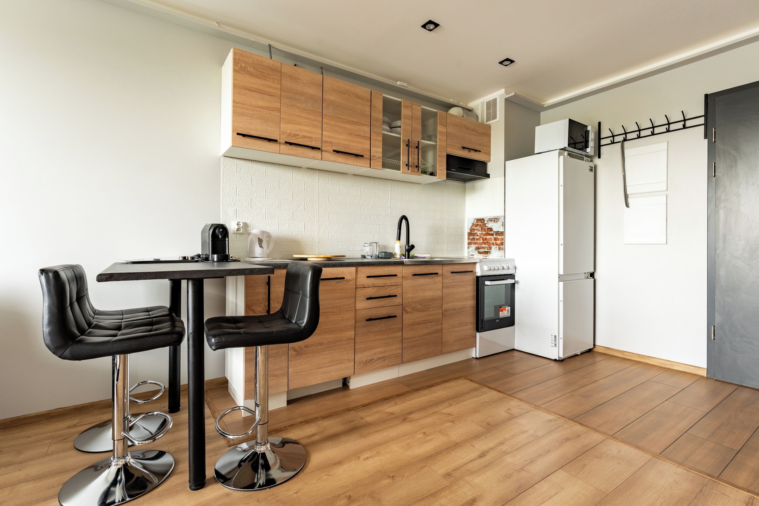 A modern kitchen with wooden cabinets, a stainless steel sink, and a white refrigerator. There are two black bar stools and a stainless steel table.