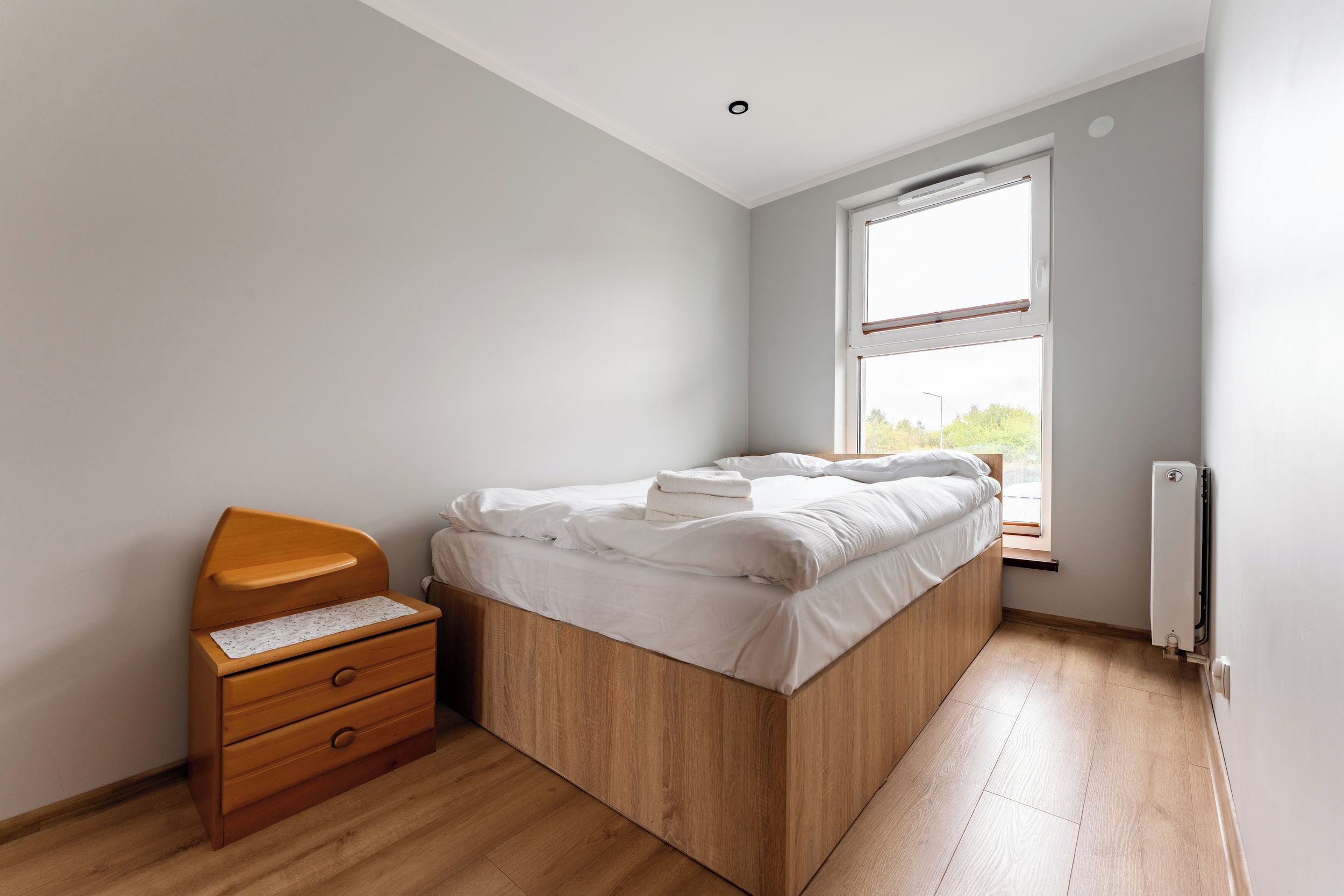 A bedroom with a wooden bed, a wooden nightstand with drawers, a window with a view, and a radiator.
