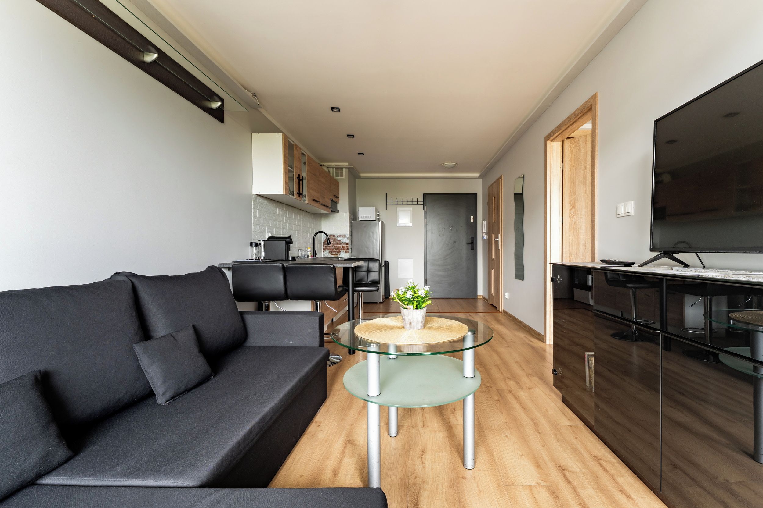 A modern living room with a dark sofa, a glass coffee table, and a kitchen area in the background.